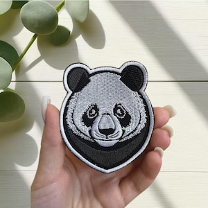 Op de afbeelding: Geborduurde panda-patch met een gedetailleerde panda-gezicht in zwart en wit. De patch heeft een zwarte rand en is ontworpen om op stof te strijken of te naaien. Het gezicht van de panda is gecentreerd, met zichtbare ogen en neus.