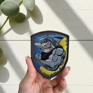 Puede incluir: Parche bordado con un tiburón musculoso flexionando su bíceps. El tiburón es gris con una sonrisa amenazante y está sobre un fondo azul y amarillo. El parche tiene forma de escudo y tiene un borde negro.