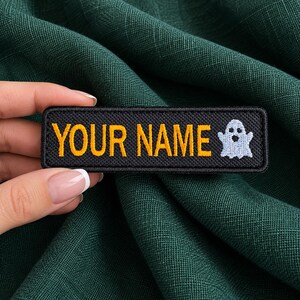Può includere: Una toppa rettangolare nera ricamata con il testo YOUR NAME in lettere maiuscole gialle, accompagnata da una piccola icona di un fantasma bianco sul lato destro. La toppa presenta un bordo cucito e una superficie in tessuto testurizzato.