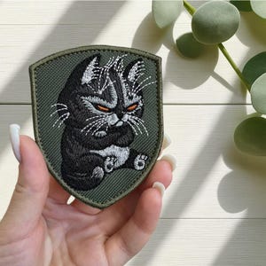 May include: Embroidered patch depicting a grumpy black cat with orange eyes and white whiskers. The cat is seated with its paws crossed. The patch is shield-shaped with a green border and backing. A fun accessory for jackets, bags, or other fabric items.