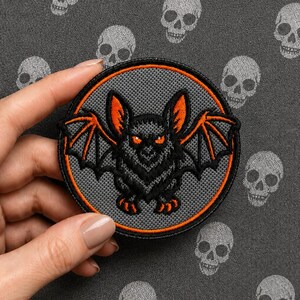 Op de afbeelding: Een ronde geborduurde patch met een zwarte vleermuis met oranje ogen, oren en vleugelaccenten. De vleermuis is gecentreerd op een grijze textuurachtergrond, omsloten door een dunne oranje rand met een zwarte gestikte randafwerking.