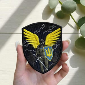 Peut inclure: Écusson brodé représentant un soldat avec des ailes jaunes, tenant une épée et un bouclier avec un symbole de trident. L'écusson est noir avec des accents jaunes et bleus. Le design comprend des éclairs.