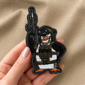 Op de afbeelding: Een geborduurde patch met een cartoonpinguïn in tactische militaire uitrusting, inclusief een helm met nachtkijker, een vest en een geweer dat rechtop wordt gehouden. De pinguïn heeft een witte buik, zwarte vleugels en oranje voeten.