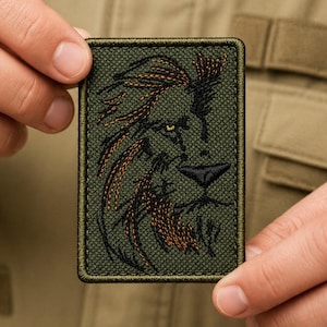 May include: A rectangular olive green embroidered patch featuring a lion's head. The lion's mane is detailed in shades of brown and orange, with black outlining the face and features. The patch is held between two hands.