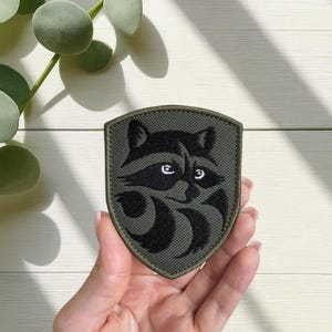 以下が含まれることがあります： アライグマのデザインが施されたシールド型のパッチ。アライグマは黒色で白い目を持ち、濃い緑色の背景に配置されています。パッチは手で持たれており、背景には明るい色の木製の表面があります。