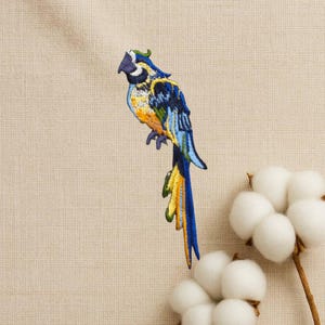 May include: Embroidered patch of a colourful parrot with blue, yellow, and green feathers. The parrot is perched on a branch, with a long tail. The patch is on a beige fabric background. Cotton branch with white cotton balls in the background.