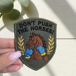 May include: Embroidered patch with a shield shape and the text "DON'T PUSH THE HORSES!". The patch features a brown horse wearing blue boxing gloves. Gold-coloured wheat stalks flank the horse on either side.