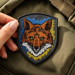 Op de afbeelding: Een schildvormige geborduurde patch met een gedetailleerde vossenkop in oranje, bruine en witte tinten. De vos is geplaatst tegen een achtergrond van blauwe en gele draden, omlijst door een dikke zwarte rand op een donkere ondergrond.