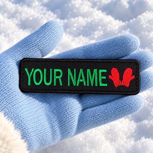 May include: A rectangular black embroidered patch featuring the text "YOUR NAME" in bright green capital letters, accompanied by a small red icon of two mittens on the right side, with a black stitched border around the entire edge.