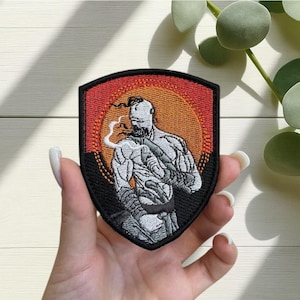 May include: Embroidered patch featuring a muscular figure with a beard and a shaved head, set against a sunset-colored background. The patch has a black border and a shield shape. The figure is depicted in shades of gray and white.