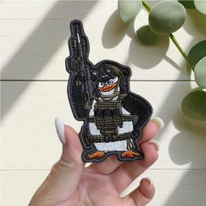 Puede incluir: Parche bordado con un pingüino de dibujos animados con equipo táctico, sosteniendo un rifle. El pingüino está representado en negro, blanco y naranja, con un chaleco y gafas de visión nocturna. El parche tiene un borde negro.
