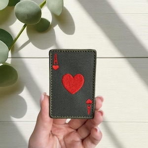 Puede incluir: Parche rectangular con el diseño de la carta As de Corazones. El parche tiene un fondo verde oscuro con un corazón rojo en el centro y símbolos 'A' rojos en las esquinas. Los bordes están cosidos con hilo verde a juego.