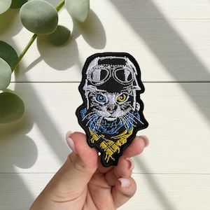 Peut inclure: Écusson brodé représentant un chat portant un casque et des lunettes d'aviateur. Le chat a les yeux bleus et jaunes, et une écharpe bleue et jaune. L'écusson est noir avec des détails blancs, bleus et jaunes.