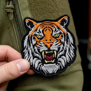 Op de afbeelding: Een geborduurde patch met de kop van een tijger met oranje, witte en zwarte vacht. De tijger heeft gele ogen, een grommende bek met zichtbare witte hoektanden en een rode tong. Het ontwerp is omlijnd met een dikke zwarte rand.
