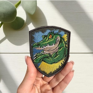 May include: Embroidered patch depicting a smiling green alligator with orange eyes. The alligator is set against a blue and yellow background, within a shield-shaped border. The patch is designed for clothing or accessory application.