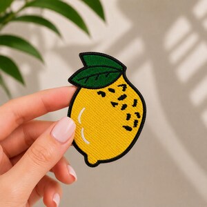 Può includere: Una toppa ricamata a forma di limone con un corpo giallo brillante, un contorno nero, una foglia verde scuro sulla parte superiore e dettagli neri testurizzati sul lato per rappresentare la buccia, rifinita con un riflesso bianco curvo.