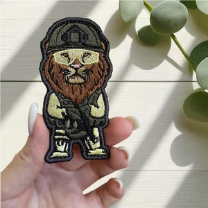 Op de afbeelding: Geborduurde patch met een leeuw in militaire kledij, compleet met helm en geweer. De patch gebruikt bruine, groene en beige draden, met een zwarte rand. Het gezicht van de leeuw is gedetailleerd en draagt een geel getinte bril.
