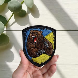 Op de afbeelding: Geborduurde patch met een bruine beer met open mond, tegen een blauw en gele achtergrond. De patch heeft een zwarte rand en is schildvormig. De beer heeft zichtbare klauwen.