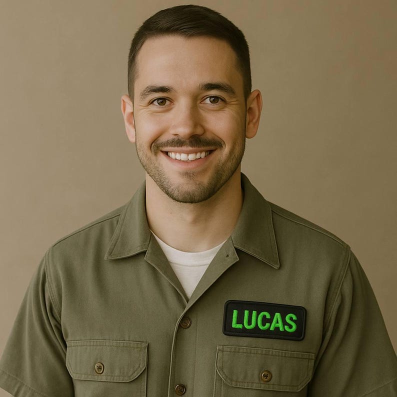K&ouml;nnte beinhalten: Eine l&auml;chelnde Person tr&auml;gt ein olivgr&uuml;nes Hemd mit einem schwarzen Aufn&auml;her mit dem Namen "LUCAS" in neongr&uuml;nen Buchstaben. Die Person hat dunkles Haar und einen Bart.