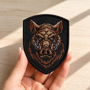 May include: A shield-shaped embroidered patch featuring a detailed brown and tan boar head design centred on a black background. The patch has a thick black stitched border and depicts the animal with sharp tusks and a focused expression.