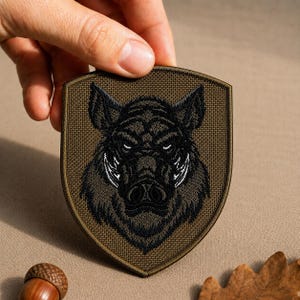 May include: A shield-shaped embroidered patch featuring a detailed black and dark gray boar head design centered on a textured olive green background. The patch has a dark olive green border and a sturdy fabric construction with visible stitching.