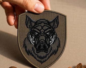 May include: A shield-shaped embroidered patch featuring a detailed black and dark gray boar head design centered on a textured olive green background. The patch has a dark olive green border and a sturdy fabric construction with visible stitching.