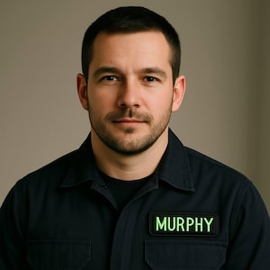 K&ouml;nnte beinhalten: Eine Person tr&auml;gt ein dunkelblaues Hemd mit Kn&ouml;pfen und einem schwarzen Rundhals-Unterhemd. Ein rechteckiger Aufn&auml;her mit dem Namen "MURPHY" in leuchtend gr&uuml;nen Buchstaben ist auf dem Hemd angebracht.