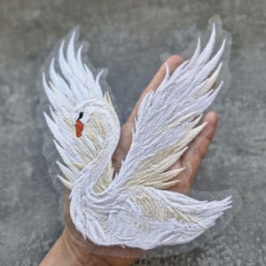 May include: Embroidered white swan patch with outstretched wings. The swan features detailed stitching in white and light beige, with a red beak and black eye. The patch is on a sheer, light-colored backing, suitable for sewing or ironing onto fabric.