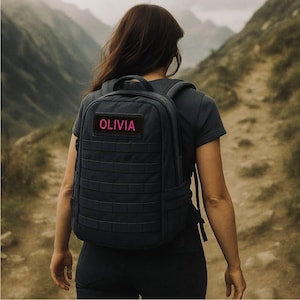K&ouml;nnte beinhalten: Ein dunkelgrauer Rucksack mit einem rechteckigen Aufn&auml;her mit dem Namen "OLIVIA" in rosa Schrift. Der Rucksack hat ein gitterartiges Design und wird auf einem Pfad mit Bergen im Hintergrund getragen.