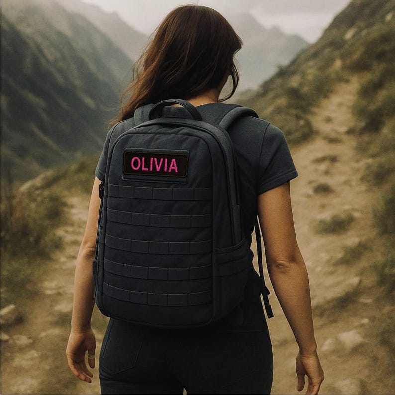 K&ouml;nnte beinhalten: Ein dunkelgrauer Rucksack mit einem rechteckigen Aufn&auml;her mit dem Namen "OLIVIA" in rosa Schrift. Der Rucksack hat ein gitterartiges Design und wird auf einem Pfad mit Bergen im Hintergrund getragen.