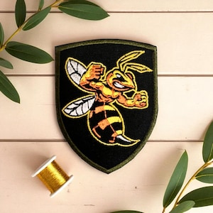 May include: Embroidered patch featuring a stylized, muscular yellow and black wasp on a black shield-shaped background. The patch has a green border and is likely for clothing or accessories. A spool of gold thread is also visible.