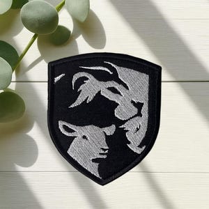 May include: Black and silver embroidered patch with a shield shape. The patch features a lamb and a lion design. The patch is likely for a jacket, backpack, or other apparel.