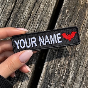 May include: A rectangular black embroidered patch featuring the white text YOUR NAME in capital letters followed by a small red bat icon. The patch has a black stitched border and is designed for customisation with a name or specific text.