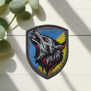 May include: Embroidered patch featuring a howling wolf with red eyes and open mouth. The wolf is gray and black, set against a blue and yellow background within a gray shield shape. The patch is likely for clothing or accessories.