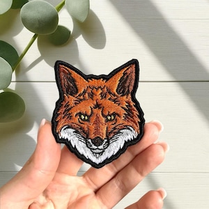 Könnte beinhalten: Gestickter Aufnäher mit einem detaillierten Fuchskopf mit orangefarbenem, braunem und weißem Fell, umrandet von einem schwarzen Rand. Der Fuchs hat gelbe Augen und einen fokussierten Ausdruck. Geeignet für Jacken, Taschen oder andere Textilien.