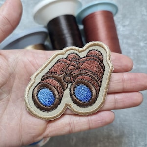 May include: Embroidered patch featuring a detailed design of binoculars. The patch has brown and blue accents, with a tan border. The binoculars are the focal point, suggesting a theme of exploration or outdoor activities.