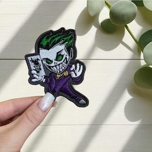 Pode incluir: Patch bordado com uma versão de desenho animado do personagem Coringa. O patch tem uma borda preta, cabelo verde e um terno roxo. O Coringa está segurando uma carta de baralho e tem um sorriso sinistro.