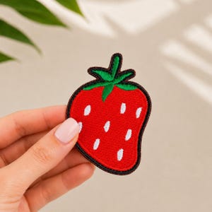 Puede incluir: Un parche bordado con forma de fresa que presenta un cuerpo de color rojo vibrante con detalles de semillas cosidas en blanco y un tallo de hojas verde brillante en la parte superior. El parche tiene un borde negro oscuro bien definido.