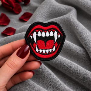 May include: An embroidered patch featuring a pair of open red lips with sharp white fangs. The design is outlined in black thread, showcasing a detailed mouth with visible teeth and a red tongue, creating a high-contrast graphic element for clothing.