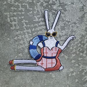 Pode incluir: Patch bordado com um coelho branco usando óculos de sol e um maiô rosa, com uma boia listrada azul e branca. O coelho está deitado, com as pernas estendidas e um braço levantado. O patch tem um design detalhado e colorido.