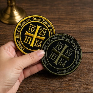 Könnte beinhalten: Zwei gestickte Aufnäher mit religiösem Text und einem Kreuzdesign. Ein Aufnäher ist schwarz mit goldfarbenen Buchstaben und einem goldenen Kreuz. Der andere ist schwarz mit olivgrünen Buchstaben und einem grünen Kreuz. Die Aufnäher sind rund.