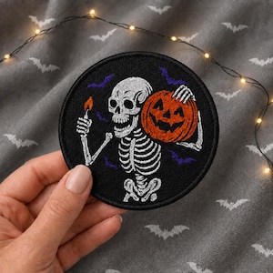 May include: A circular embroidered patch featuring a white skeleton on a black background. The skeleton holds a small orange pumpkin in one hand and a lit match with a bright orange flame in the other. Small purple bats are scattered around the figure.