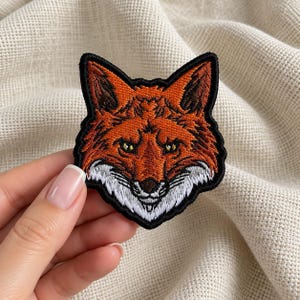 Könnte beinhalten: Ein gestickter Aufnäher mit dem Kopf eines Fuchses, gearbeitet mit orangefarbenen, braunen und weißen Garnen. Das Motiv ist von einem dicken schwarzen Rand umgeben und zeigt die spitzen Ohren, die wachen Augen und das strukturierte Fell.