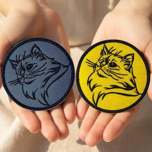 Può includere: Due toppe rotonde a forma di gatto. Una è blu navy con un disegno di gatto nero, l'altra è gialla con un disegno di gatto nero. Entrambe hanno un bordo nero e sono tenute in mani aperte.