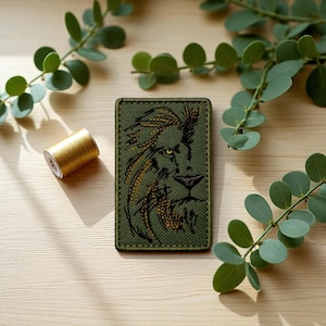 Może przedstawiać: Oliwkowozielona prostokątna naszywka z haftowaną głową lwa w kolorze złotym i czarnym. Po lewej stronie znajduje się szpula złotych nici, a w tle zielone liście. Naszywka jest prawdopodobnie przeznaczona na odzież lub akcesoria.