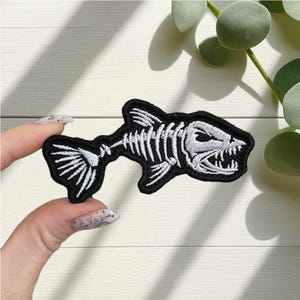May include: An embroidered patch with a fish skeleton design. The patch has a black border and the fish skeleton is white. The fish has visible teeth and a detailed tail. This patch is suitable for jackets, bags, or other fabric items.