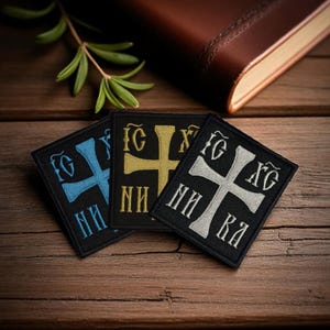 Puede incluir: Tres parches bordados con símbolos religiosos. Cada parche es negro con un diseño de cruz en azul, dorado y plateado. Los parches son rectangulares y presentan texto cirílico. Un libro encuadernado en cuero y vegetación en el fondo.