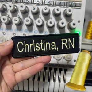 K&ouml;nnte beinhalten: Ein schwarzer, bestickter Aufn&auml;her mit dem Namen "Christina, RN" in gelbem Faden. Der rechteckige Aufn&auml;her wird in einer Hand gehalten, mit einer N&auml;hmaschine und einer gelben Garnrolle im Hintergrund.