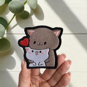May include: Embroidered patch showing two cartoon cats. A brown cat embraces a light blue cat holding a red heart. The patch has a black border. The image is on a light wood grain surface.
