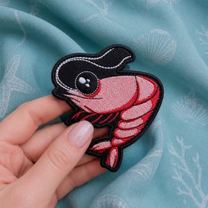 以下が含まれることがあります： 黒い頭部と白いハイライトが入った大きな目を持つ、デフォルメされたピンク色のエビの刺繍ワッペン。デザインは黒い糸で縁取りされており、湾曲した体型、節のあるディテール、そして下部には小さな尾が表現されています。